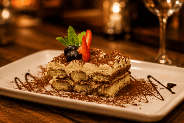 A slice of tiramisu on a white plate, topped with fresh blueberries, strawberry slices, and mint leaves, dusted with cocoa powder.