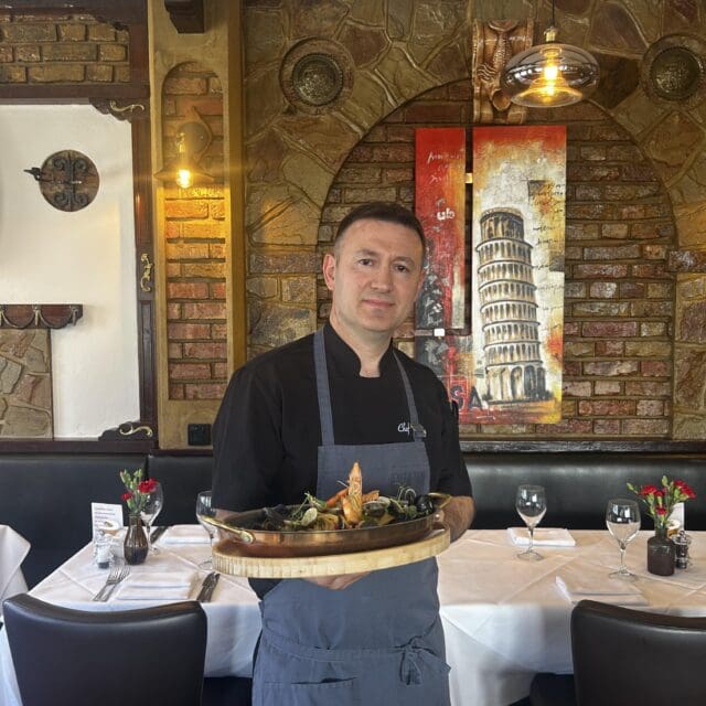 A chef in a black uniform and grey apron holds a copper pan of seafood in a restaurant with brick walls.